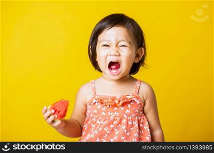 Happy portrait Asian baby or kid cute little girl attractive laugh smile wearing t-shirt playing holds cut watermelon fresh for eating, studio shot isolated on yellow background, healthy food and summer concept
