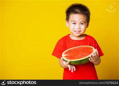Happy portrait Asian child or kid cute little boy attractive laugh smile wearing red t-shirt playing holds cut half watermelon, studio shot isolated on yellow background