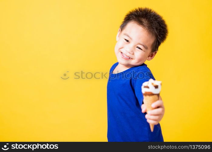 Happy portrait Asian child or kid cute little boy attractive laugh smile playing holds and eating sweet chocolate ice cream waffle cone, studio shot isolated on yellow background, summer concept