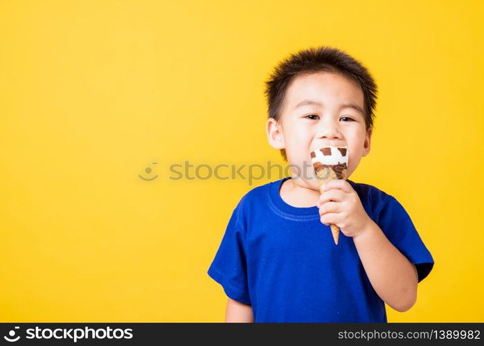 Happy portrait Asian child or kid cute little boy attractive laugh smile playing holds and eating sweet chocolate ice cream waffle cone, studio shot isolated on yellow background, summer concept