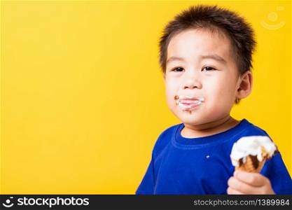 Happy portrait Asian child or kid cute little boy attractive laugh smile playing holds and eating sweet chocolate ice cream waffle cone, studio shot isolated on yellow background, summer concept