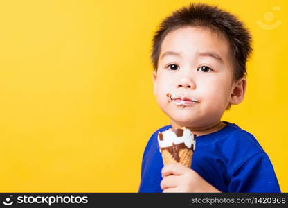 Happy portrait Asian child or kid cute little boy attractive laugh smile playing holds and eating sweet chocolate ice cream waffle cone, studio shot isolated on yellow background, summer concept