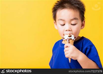 Happy portrait Asian child or kid cute little boy attractive laugh smile playing holds and eating sweet chocolate ice cream waffle cone, studio shot isolated on yellow background, summer concept