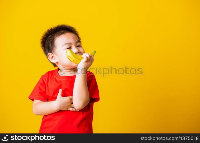 Happy portrait Asian child or kid cute little boy attractive smile wearing red t-shirt playing holds banana fruit, studio shot isolated on yellow background