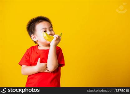 Happy portrait Asian child or kid cute little boy attractive smile wearing red t-shirt playing holds banana fruit, studio shot isolated on yellow background