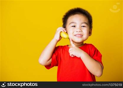 Happy portrait Asian child or kid cute little boy attractive smile wearing red t-shirt playing holds banana fruit pretending to be like a telephone, studio shot isolated on yellow background