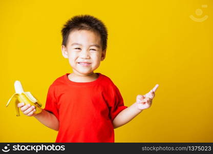 Happy portrait Asian child or kid cute little boy attractive smile wearing red t-shirt playing holds peeled banana for eating, studio shot isolated on yellow background