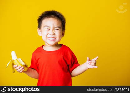 Happy portrait Asian child or kid cute little boy attractive smile wearing red t-shirt playing holds peeled banana for eating, studio shot isolated on yellow background