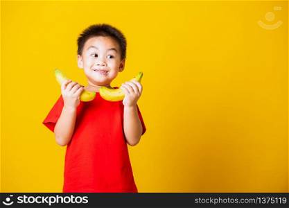 Happy portrait Asian child or kid cute little boy attractive smile wearing red t-shirt playing holds banana fruit, studio shot isolated on yellow background