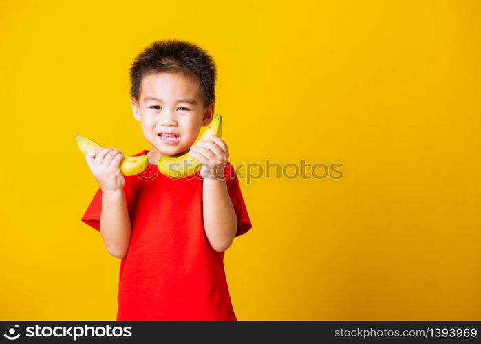 Happy portrait Asian child or kid cute little boy attractive smile wearing red t-shirt playing holds banana fruit, studio shot isolated on yellow background