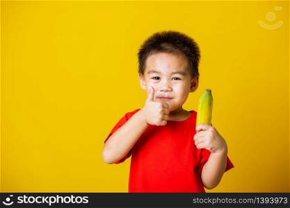Happy portrait Asian child or kid cute little boy attractive smile wearing red t-shirt playing holds bananas and show finger thumb for good sign, studio shot isolated on yellow background