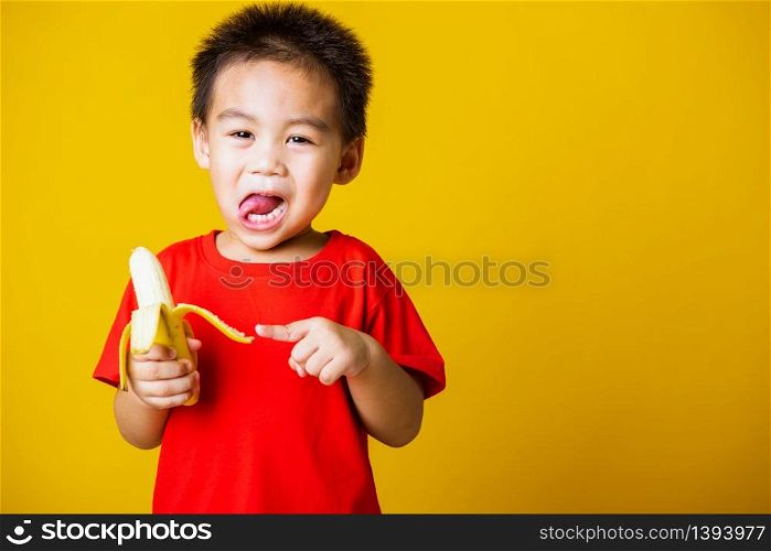 Happy portrait Asian child or kid cute little boy attractive smile wearing red t-shirt playing holds peeled banana for eating, studio shot isolated on yellow background