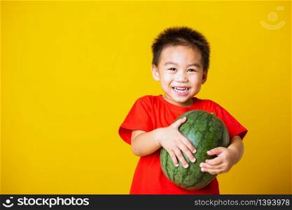 Happy portrait Asian child or kid cute little boy attractive smile wearing red t-shirt playing holds full watermelon that has not been cut, studio shot isolated on yellow background