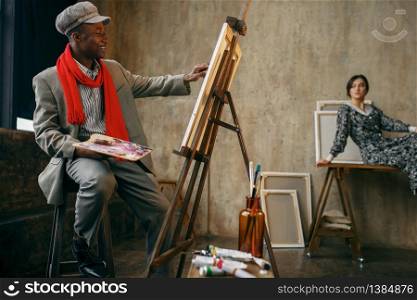 Happy portrait painter with brush draws female model in art studio. Male artist at his workplace, creative master at the easel in workshop. Happy painter draws female model in art studio