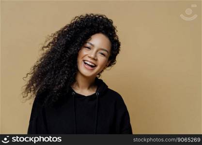 Happy positive African American woman with curly hair, tilts head and smiles broadly, dressed in casual black sweatshirt, isolated on beige background, expresses positive emotions. People, happiness