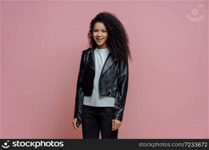 Happy positive Afro American woman dressed in leather jacket, ready for outdoor walk, poses against pink background. Joyful dark skinned millennial girl in good mood has casual talk with friend