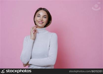 Happy positive young European woman stands confident smiles pleasantly, keeps hand near face, poses against pink background, expresses positive emotions. People, lifestyle and beauty concept
