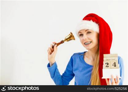 Happy positive young teenage woman wearing Santa Claus christmas hat holding calendar with 24 december date and bell. Woman and christmas calendar with bell