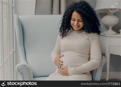 Happy pregnancy. Pretty mixed race woman with curly hair in white casual dress holding her belly in anticipation of future baby, thinking about maternity and how to be caring and loving mother. Pretty african american woman holding her belly in anticipation of future baby