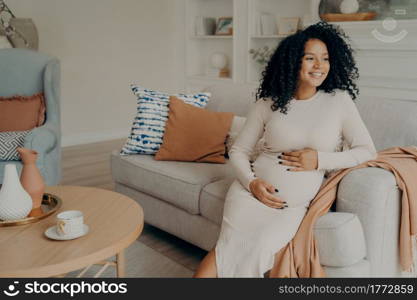 Happy pregnant afro american woman in casual dress with curly hair looking at window while sitting on couch in living room, checking out weather and thinking about going outside for walk. Pregnant afro american woman looking at window while sitting on sofa at home