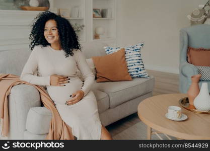 Happy pregnant afro american woman in casual dress with curly hair looking at window while sitting on couch in living room, checking out weather and thinking about going outside for walk. Pregnant afro american woman looking at window while sitting on sofa at home