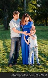 Happy pregnant family of three expecting new baby. Father and mother with little boy hugging outdoors.