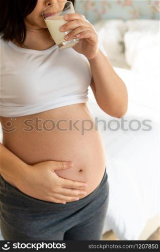 Happy pregnant woman drinks milk in glass at home while taking care of her child. The young expecting mother holding baby in pregnant belly. Calcium food nutrition for strong bones of pregnancy.