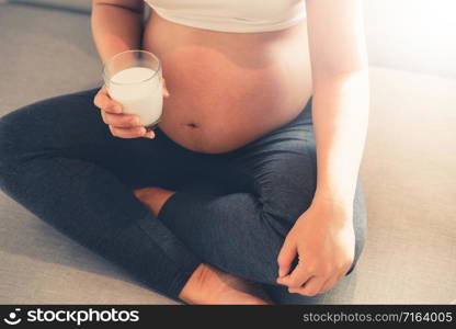 Happy pregnant woman drinks milk in glass at home while taking care of her child. The young expecting mother holding baby in pregnant belly. Calcium food nutrition for strong bones of pregnancy.