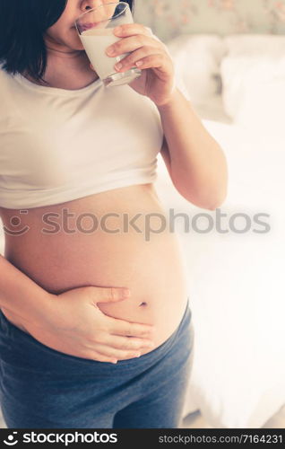 Happy pregnant woman drinks milk in glass at home while taking care of her child. The young expecting mother holding baby in pregnant belly. Calcium food nutrition for strong bones of pregnancy.