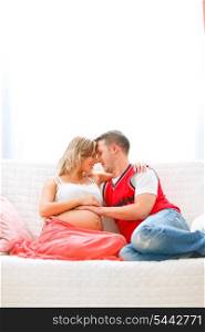 Happy pregnant woman sitting with husband on couch and hugging&#xA;