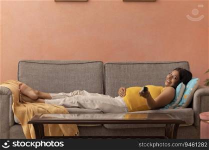 Happy pregnant woman watching TV while relaxing on sofa at home