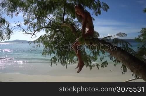 Happy pretty blonde woman wearing colourful swimwear sitting on a branch on a beautiful summer day with beautiful sea and sky background - video in slow motion