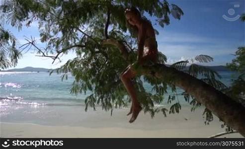 Happy pretty blonde woman wearing colourful swimwear sitting on a branch on a beautiful summer day with beautiful sea and sky background - video in slow motion