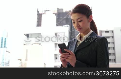Happy pretty Japanese female manager using phone. Portrait of young businesswoman, successful girl, busy woman at work, texting with smartphone, text messaging mobile telephone near office buildings