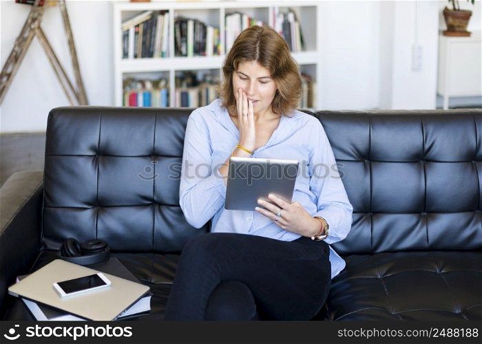Happy pretty young woman in casual outfit sitting on sofa at home and using tablet