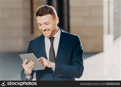 Happy prosperous businessman in formal wear, makes money transaction via touch pad, has glad expression, stands outdoor, connected to wireless intrnet. People, business and technology concept