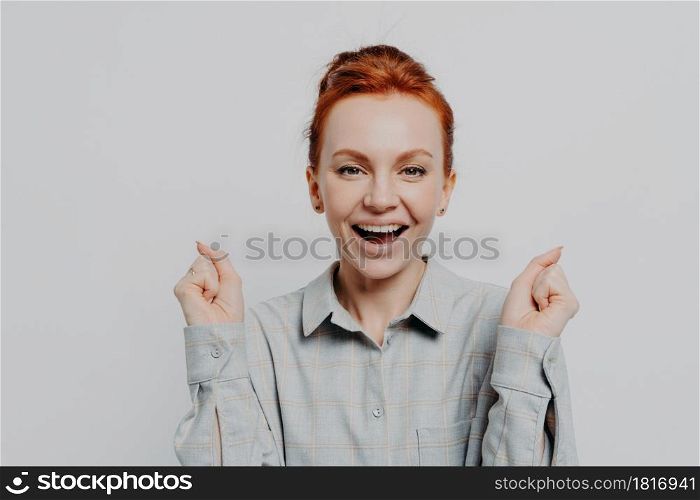 Happy red haired 30s woman wearing casual shirt holding clenched fists and looking at camera with overjoyed emotion, enjoying moment of success and celebrating victory, isolated on grey background. Happy excited red haired woman holding clenched fists and celebrating success in studio