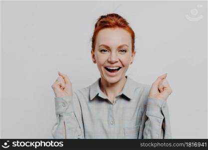 Happy red haired 30s woman wearing casual shirt holding clenched fists and looking at camera with overjoyed emotion, enjoying moment of success and celebrating victory, isolated on grey background. Happy excited red haired woman holding clenched fists and celebrating success in studio