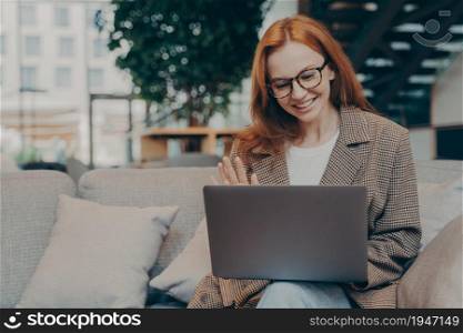 Happy red-haired business woman in optical eyewear waving at laptop webcam while sitting on sofa and having web conference with client, engaged in video call with colleagues while working remotely. Happy red-haired business woman waving at laptop webcam during web conference with client