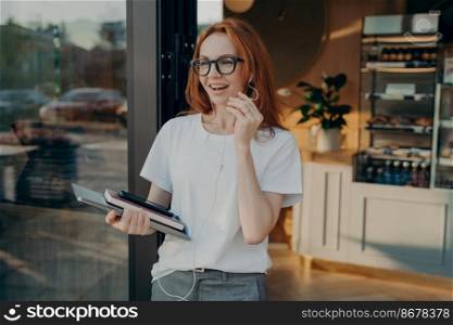 Happy red-haired businesswoman with laptop in hand having call on mobile phone, leaving coffee shop, smiling female in eyeglasses using earphones and smartphone, talking with partner, standing outdoor. Happy red-haired businesswoman with laptop in hand having call on mobile phone, leaving coffee shop