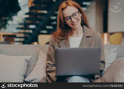 Happy red-haired female office worker chatting on laptop computer while sitting on grey couch with lots of pillows, comfortable office break room, business woman relaxing in her spare time at work. Happy female office worker chatting on laptop while sitting on grey couch at office