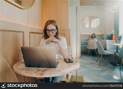 Happy redhaired woman has distance job works on laptop computer in cozy cafe listens webinar online while having coffee watches tutorial video. Intelligent student prepares for exams in coffee shop. Happy redhaired woman has distance job works on laptop computer in cozy cafe