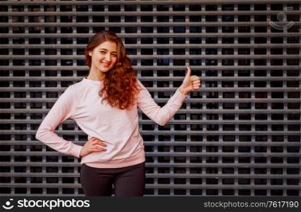Happy redhead girl shows us thumb up sign in front of metal grid. Happy redhead girl shows us thumb up sign