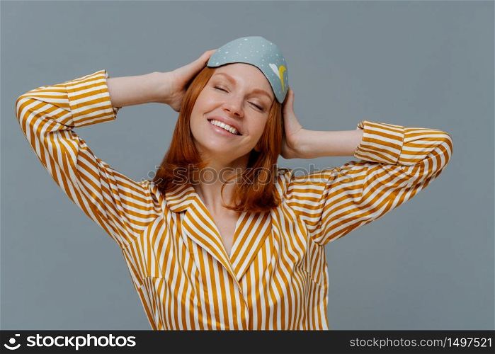 Happy redhead young woman had enough sleep, keeps hands on head, stands with closed eyes, wears cozy homewear and blindfold, isolated over grey background, has good rest. Sleeping time concept