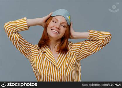 Happy redhead young woman had enough sleep, keeps hands on head, stands with closed eyes, wears cozy homewear and blindfold, isolated over grey background, has good rest. Sleeping time concept