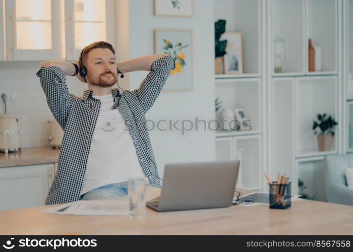 Happy relaxed bearded man holding hands behind head and pausing while working online from home using headset and laptop, male freelancer resting after hard day. Remote job and freelance concept. Calm young freelancer resting while sitting at his workplace at home