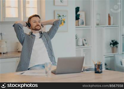 Happy relaxed bearded man holding hands behind head and pausing while working online from home using headset and laptop, male freelancer resting after hard day. Remote job and freelance concept. Calm young freelancer resting while sitting at his workplace at home