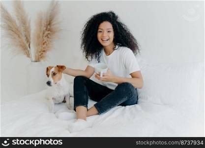 Happy relaxed lovely young woman with Afro hairstyle, petting dog, sit together on bed with white bedclothes, drink hot beverage, dressed in casual wear, expresses positive emotions. People and rest
