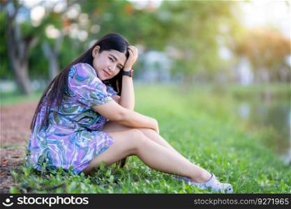 Happy Relaxing Portrait asian woman Wear purple dress wearing smartwatch while sitting on green grass lawn beside a reservoir at the city park outdoors.