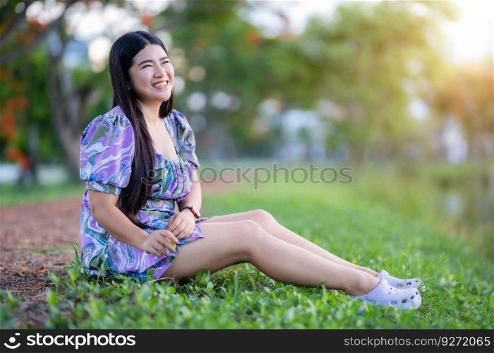Happy Relaxing Portrait asian woman Wear purple dress wearing smartwatch while sitting on green grass lawn beside a reservoir at the city park outdoors.